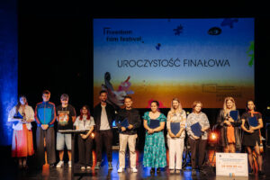 Grupa laureatów i organizatorów stoi na scenie podczas gali finałowej Freedom Film Festival. W tle znajduje się kolorowy ekran z napisem 'Uroczystość Finałowa' oraz logotypami festiwalu i programu Kameralne Lato. Uczestnicy trzymają dyplomy i nagrody, a po prawej stronie znajduje się symboliczny czek o wartości 10 000 zł za Grand Prix konkursu filmowego.