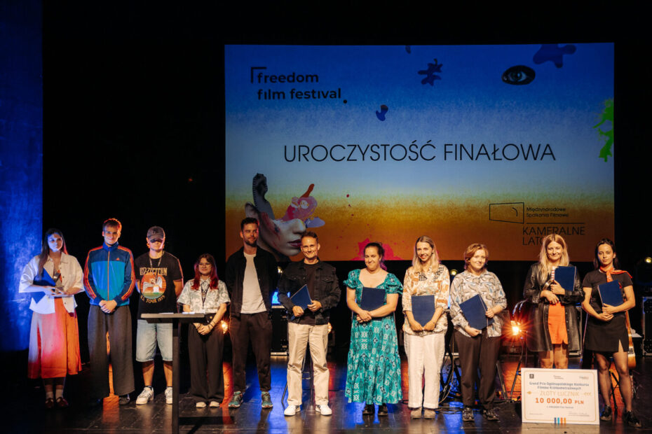 Grupa laureatów i organizatorów stoi na scenie podczas gali finałowej Freedom Film Festival. W tle znajduje się kolorowy ekran z napisem 'Uroczystość Finałowa' oraz logotypami festiwalu i programu Kameralne Lato. Uczestnicy trzymają dyplomy i nagrody, a po prawej stronie znajduje się symboliczny czek o wartości 10 000 zł za Grand Prix konkursu filmowego.