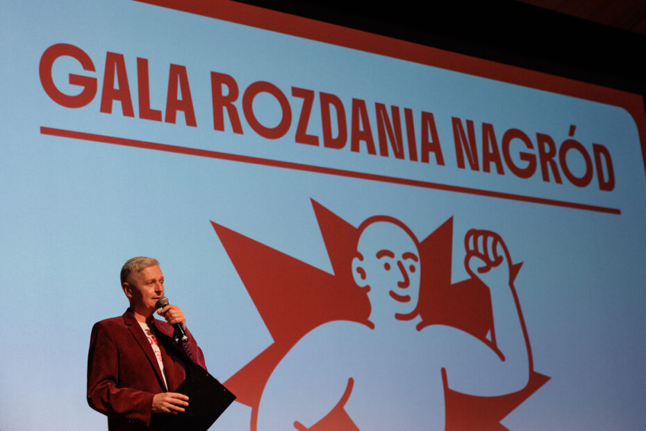 Prowadzący na scenie podczas gali rozdania nagród 17. Solanin Film Festiwal. W tle ekran z grafiką przedstawiającą rysunek statuetki i napisem "GALA ROZDANIA NAGRÓD".