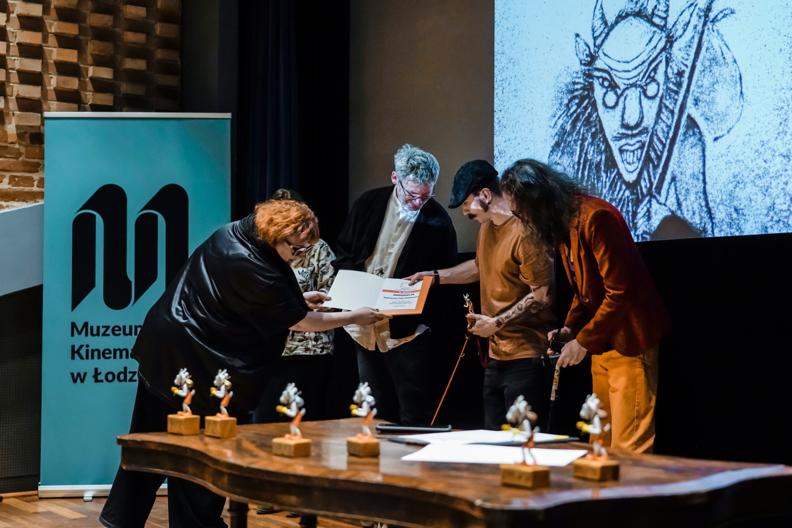 Wręczenie nominacji do Nagrody im. Jana Machulskiego podczas finału Festiwalu O!PLA w Muzeum Kinematografii w Łodzi. Kilka osób stoi przy stole ze statuetkami Koziołka Matołka, odbierając dyplom z nominacją.