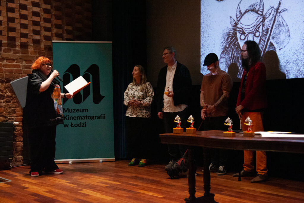 Osoby nominowane do Nagrody im. Jana Machulskiego stoją na scenie podczas finału Festiwalu O!PLA w Muzeum Kinematografii w Łodzi. Kuratorka Nagród odczytuje nominacje, na pierwszym planie widać statuetki Koziołka Matołka.